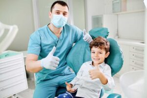A parent helps a child brush teeth with a cup and toothbrush on a bathroom sink.