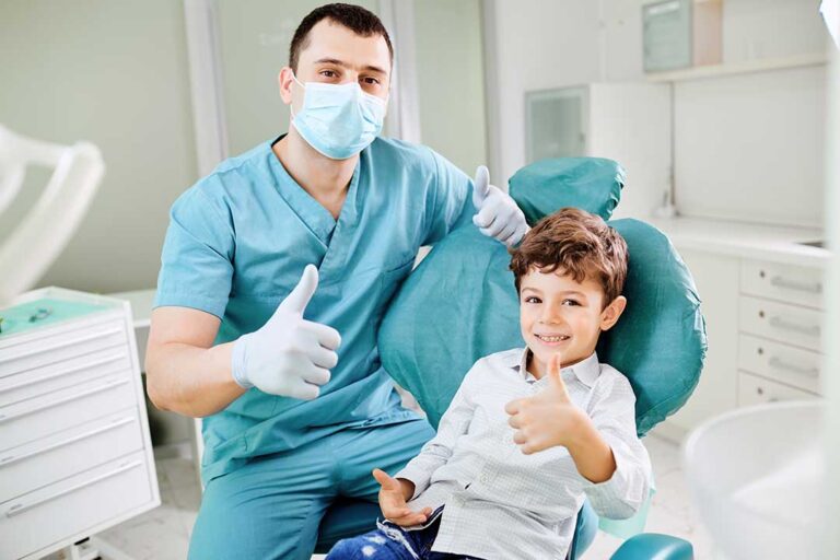 A parent helps a child brush teeth with a cup and toothbrush on a bathroom sink.